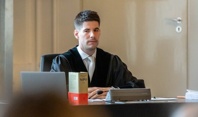 Richter Heim bleibt skeptisch. Er verweist auf die Verhältnismäßigkeit, die bei Hausdurchsuchungen gewahrt bleiben müsse und erklärt, dass die Privatsphäre eines Beschuldigten auch bei einer Razzia nicht vollständig außer Kraft gesetzt ist. Das bedeute auch, dass beim Fotografieren in einer Privatwohnung nicht alles erlaubt ist. "Das höre ich so zum ersten Mal", murmelt der Beamte S. verdutzt. Er war verantwortlich dafür, eine Lichtbildmappe zum Einsatz anzufertigen und ging bei der Bildauswahl offenbar recht großzügig vor. Abgelichtet wurden auch die Toilette und der Kleiderschrank des Beschuldigten. S. betont, die Aufnahmen nicht selbst angefertigt zu haben habe sie aber der Akte beigefügt. Welchen Erkenntnisgewinn er sich davon versprach und wo er den Beweiswert sieht? Der Kriminalbeamte erzählt frei von der Leber weg, er sei der Ansicht, der Zustand einer Wohnung helfe dabei, eine Person zu beurteilen und eine ordentliche Haushaltsführung lasse seiner Erfahrung nach auf verlässlichere Angaben schließen.  Da wird der Vorsitzende Richter deutlich: Nach einer groben und vorläufigen Einschätzung halte er die Mehrzahl der Bilder in der Mappe für rechtswidrig, nur 9 von 26 seien legitim. Für Heim ist auch nicht nachvollziehbar, wozu es eine Skizze vom Grundriss der Wohnung gebraucht hat, das kennt er vielleicht vom bewaffneten Handel mit Rauschgift, aber doch nicht, um eine Urheberschaft festzustellen, die schon eingeräumt war. S. rutscht nervös auf dem Stuhl herum, schaut sich hilfesuchend um – da springt ihm Staatsanwalt Graulich bei, der die Razzia veranlasst hat: "Zur Wahrheit gehört auch, dass das OLG Stuttgart die Rechtmäßigkeit der Hausdurchsuchung festgestellt hat.". Richter Heim: "Vielleicht die Anordnung, aber nicht die Art und Weise der Durchführung." Graulich: "Doch." Heim: "Nein." Schmollend verschränkt der Staatsanwalt die Arme und bedankt sich beim Zeugen noch einmal recht herzlich für die gute Ermittlungsarbeit.  Zwei Gerichte, zwei divergierende Rechtsauffassungen Die Vorgeschichte zum Prozess verdeutlicht ganz gut, warum es heißt, vor Gericht und auf hoher See lägen Schicksale in Gottes Hand. Denn der Fall macht deutlich, wie unterschiedlich derselbe Sachverhalt von verschiedenen Richter:innen bewertet werden kann. Weil Kienert einen Link gesetzt hat, wird von der deutschen Justiz aufwändig überprüft, ob er sich strafbar gemacht hat. In Frage kommt zum Beispiel § 85 des Strafgesetzbuches: Falls festgestellt werden sollte, dass der Journalist mit seiner Handlung die Fortführung einer verbotenen Vereinigung unterstützt hat, drohen ihm eine Geldstrafe oder bis zu drei Jahre Haft.